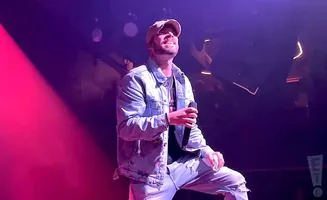 A man wearing a denim jacket and baseball cap sings into a microphone on a stage with purple and pink lighting.