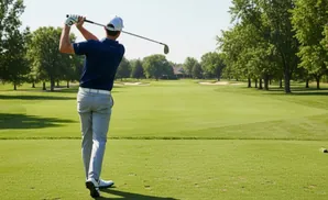 Golfer in mid-swing on a sunny golf course with trees and a clubhouse in the background.