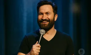 A smiling man with a beard holds a microphone while wearing a black V-neck shirt, with a blue curtain in the background.