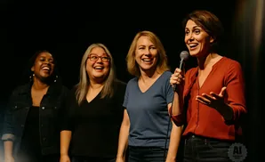 Four women laugh on stage, one holding a microphone.