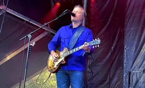 A man in a blue shirt plays a guitar and sings into a microphone on stage.