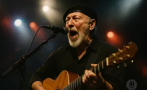 A man in a black beret and shirt sings into a microphone while playing an acoustic guitar on stage.