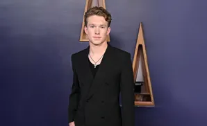 A young man in a black suit and a silver cross necklace poses against a purple wall with triangular wooden decor.