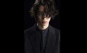 Man with dark, messy hair and a black suit jacket over a black shirt, looking forward against a black background.