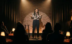 A female comedian performs on a brick stage under spotlights to a dark audience.