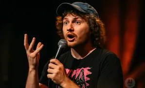 A man wearing a baseball cap and t-shirt holds a microphone and gestures with his hand.