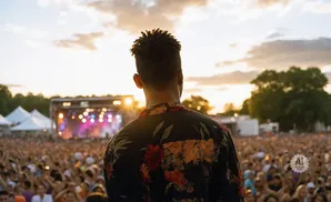 A person with dreadlocks faces away from the camera, wearing a floral shirt at an outdoor concert with a large crowd.