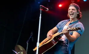A male guitarist in a blue shirt and jeans sings passionately into a microphone on stage.