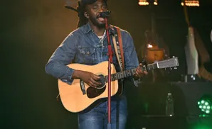 A Black man with dreadlocks wears a denim jacket and hat while playing an acoustic guitar on stage.