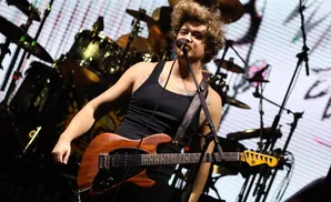 A male singer with wild hair plays a brown electric guitar during a live concert performance.