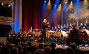 A conductor in a tuxedo smiles and directs an orchestra with a baton and raised finger.