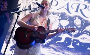 Woman with blonde hair plays guitar and sings into a microphone on stage with a blue and white patterned backdrop.