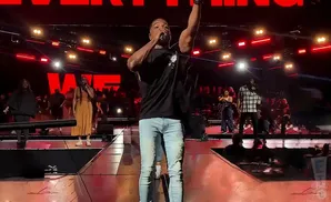 A male singer with his arm raised, performing on stage with a choir behind him.