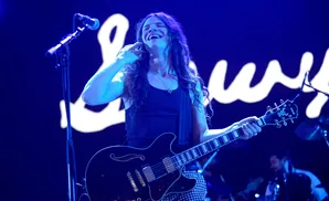 A male guitarist with long hair laughs on stage, holding a black Gibson guitar.