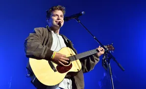 A man plays an acoustic guitar and sings into a microphone on a blue stage.