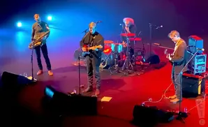 Musicians perform on a stage with blue and red lights. A saxophonist, guitarist, and drummer are visible.