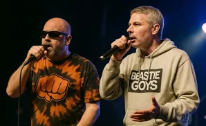Two men with microphones on stage, one wearing sunglasses and a tie-dye shirt, the other a gray hoodie with 