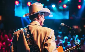 A cowboy on stage with a guitar and microphone, back to the camera, under colorful stage lights.