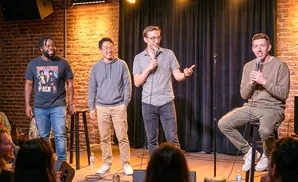 Four men on a comedy stage, three holding microphones, with brick walls and a black curtain behind them.