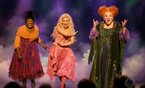 Three women in witch costumes perform on stage with purple and blue lighting.