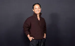 A man smiles at the camera, hands in his pockets, wearing a dark brown sweater and black pants.