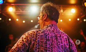 Man in a patterned shirt on a stage with bright lights and a blurred audience.