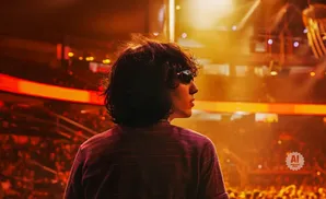 A person with dark, curly hair and sunglasses looks to the right at a concert with bright stage lights.