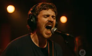 A man with curly hair sings into a microphone while wearing headphones.