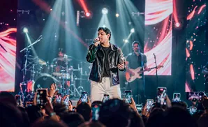 Singer on stage with a band, performing for an audience holding up phones.