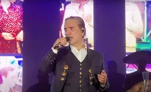 A man in a suit sings into a microphone on a stage with a colorful screen behind him.