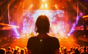 Woman silhouetted against bright stage lights at a concert, facing a large screen.