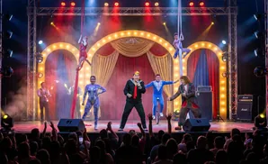 Circus performers on stage: aerialists, juggler, unicyclist, singer, and clown entertain an audience.