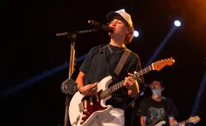 A young guitarist sings into a microphone while playing a white electric guitar on a dark stage.