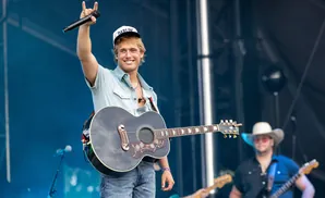 A male country singer with a guitar on stage holds a microphone and wears a baseball cap.