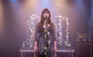 A woman in a flowing gray dress sings into a microphone adorned with white flowers and lights.