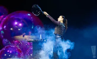 A performer creates a large bubble with a wand surrounded by purple spheres and blue smoke on a stage.