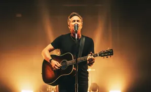 A man sings into a microphone while playing an acoustic guitar on a dimly lit stage.