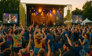 An audience cheers with hands raised at an outdoor music festival with a band performing on stage.