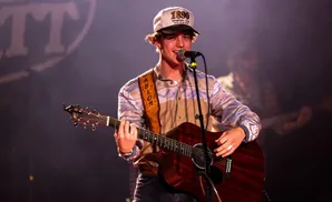 A young man with curly brown hair and a baseball cap sings and plays an acoustic guitar on stage.