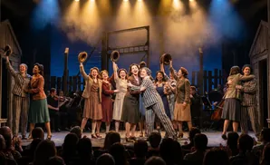 A group of women in striped uniforms and dresses on stage, holding hats in the air.