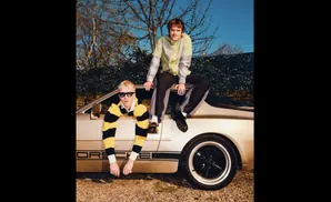 Two men pose by a Porsche. One leans out the window, the other sits on the roof.