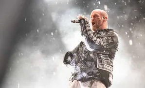 A bald man with tattoos on his face sings into a microphone on a smoky stage.