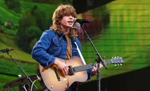 Man with long hair wearing a denim jacket plays an acoustic guitar and sings into a microphone at an outdoor concert.
