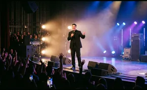 A priest in a suit on stage speaks into a microphone, with a crowd in the foreground and stage lights and equipment behind him.