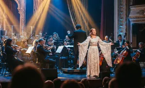 A female singer in a white dress performs with an orchestra on stage, lit by spotlights.