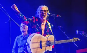Musician singing and playing a 12-string guitar on stage with blue lighting.