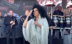 Diana Ross sings on stage in a sparkly gown, accompanied by a band with horns and percussion.