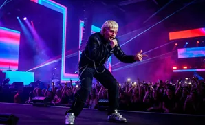 A blonde musician in pink sunglasses and a black jacket performs on stage with a crowd in the background.