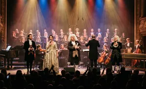 A choir and orchestra perform on stage, with solo singers in period costumes at the front.