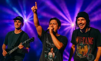 Three men perform on stage with purple stage lights; two are singing into microphones and one plays guitar.